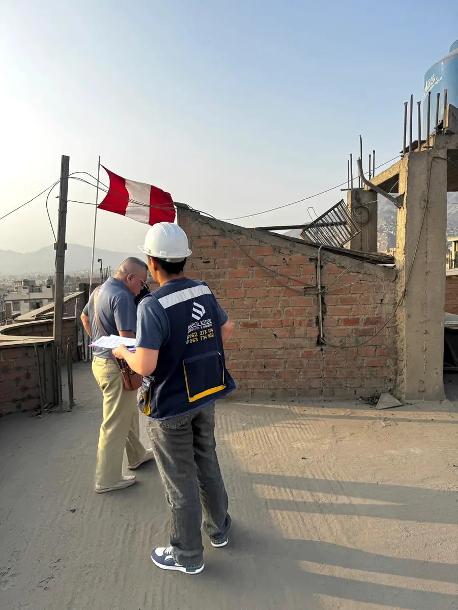 Profesionales con casco revisando documentos junto a un muro y la bandera del Perú